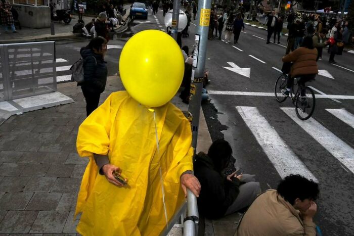 Person in yellow raincoat with yellow balloon on busy Israeli street by Ilan Ben Yehuda.