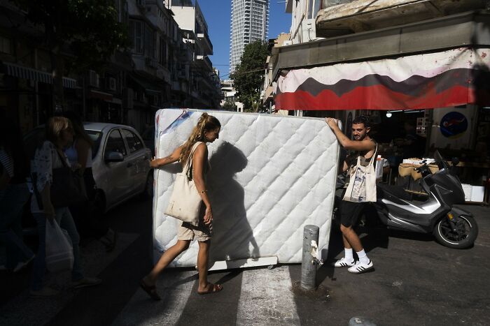 People moving a mattress on a busy street in Israel, with shops and scooters in the background.