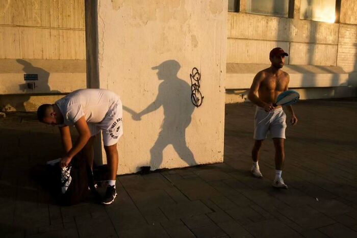 Street scene in Israel with two men, one packing a bag and another holding a paddle, shadow cast on a wall.