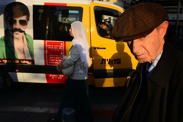 Street scene in Israel showing a man in a cap and a woman walking past a yellow van with an advertisement.