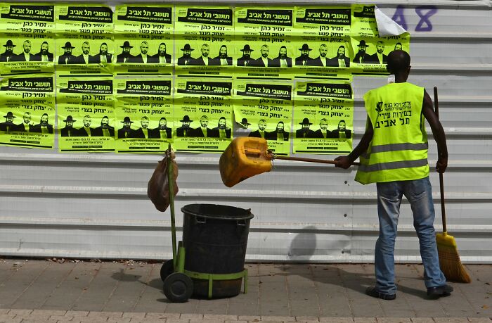 Street cleaner in a yellow vest sweeping in front of vibrant posters, Israel street photo by Ilan Ben Yehuda.