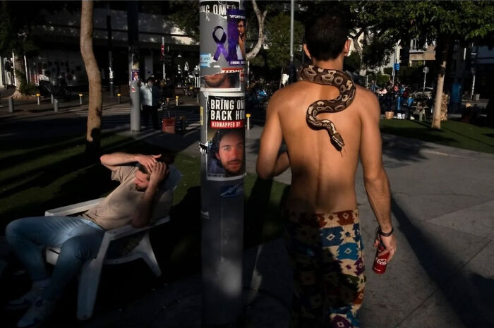 Bare-chested man with a snake in an Israeli street scene by Ilan Ben Yehuda, while another man relaxes on a chair nearby.