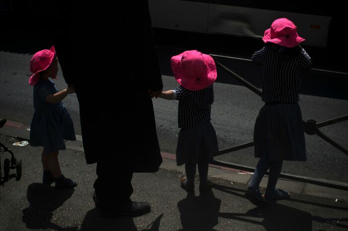 Children in pink hats holding hands with an adult, captured by Ilan Ben Yehuda, illustrating street life in Israel.