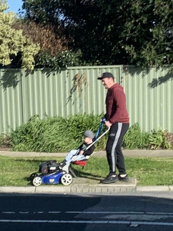 A man pushes a stroller attached to a lawnmower, with a toddler sitting calmly, depicting an unusual parenting moment.