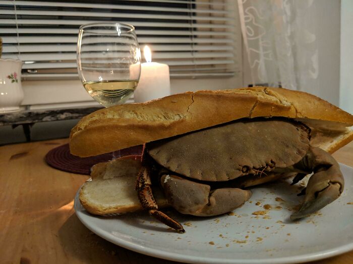 Unhinged food: A whole crab between baguette slices on a dinner plate with a glass of white wine and a lit candle.