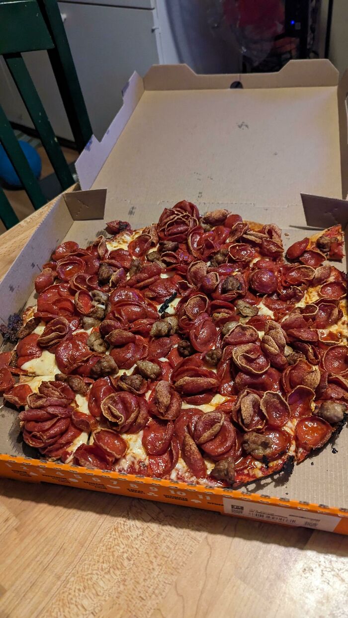 Unhinged food: a pizza overloaded with pepperoni, baked to a crisp, and presented in an open pizza box on a wooden table.