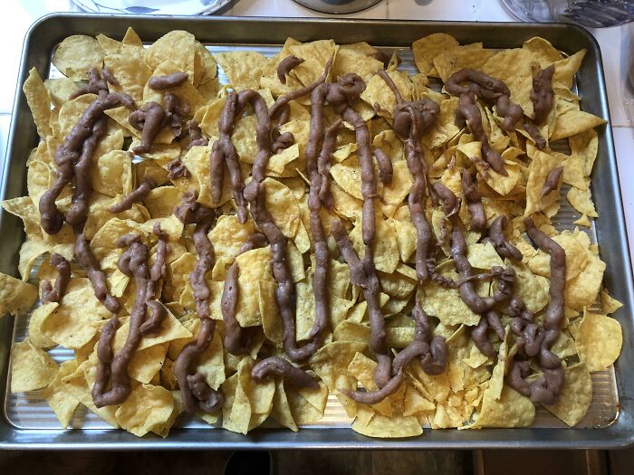 Unhinged food: Tortilla chips topped with a messy drizzle of beans on a baking tray.