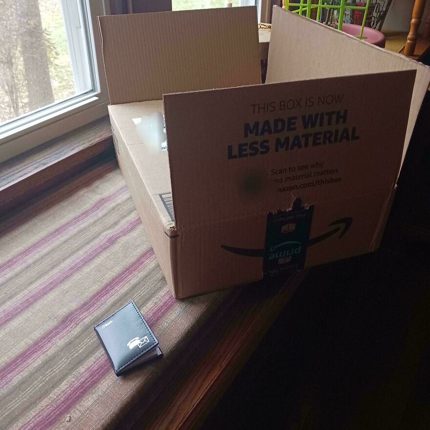 Open cardboard box labeled "Made with Less Material" beside a small wallet, illustrating wasteful packaging.