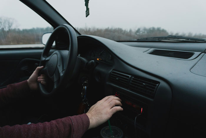 Driver adjusts car radio while holding the steering wheel, interior view during overcast weather.