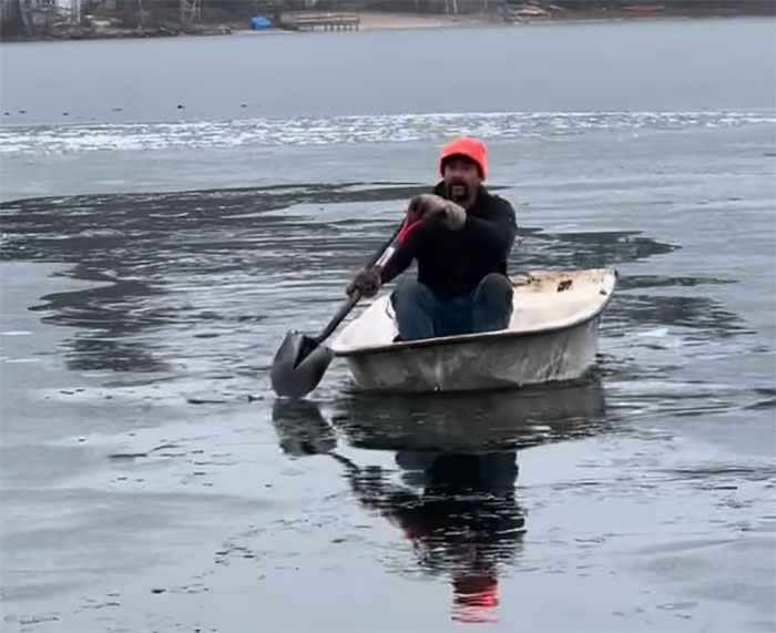 Man in a boat on icy lake rescuing a cat stuck on an ice chunk.