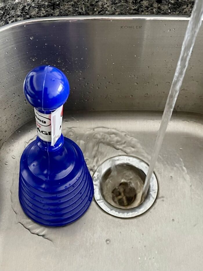 Blue sink plunger next to a running tap in a stainless steel sink.