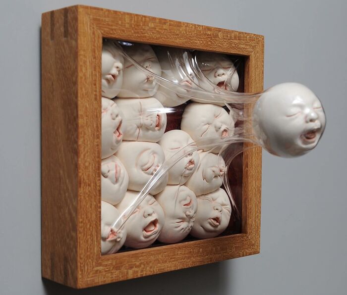 Sculpture blending realism and surrealism, featuring expressive faces in a wooden frame with one face extending outward.