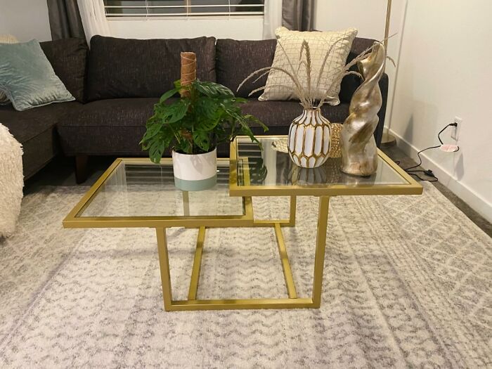 Modern glass coffee table with gold frame, decorative vases, and plant on a textured rug in a cozy living room.