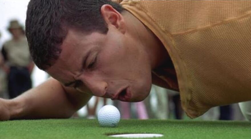 Actor Adam Sandler lying on a golf course, intently focused on a golf ball near the hole.