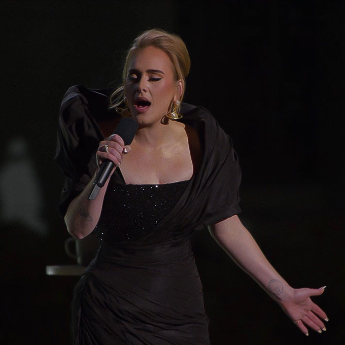 Adele performing passionately on stage in an elegant black gown, holding a microphone.