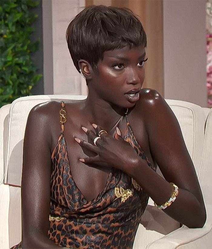 Anok Yai, a black contestant in leopard print dress, seated, expressing surprise.