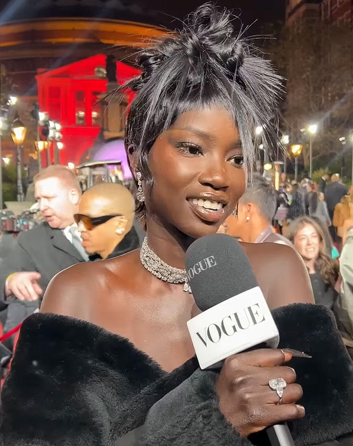 Anok Yai, a black contestant speaking at a British Fashion Awards, holding a Vogue microphone, wearing elegant jewelry and a fur stole.