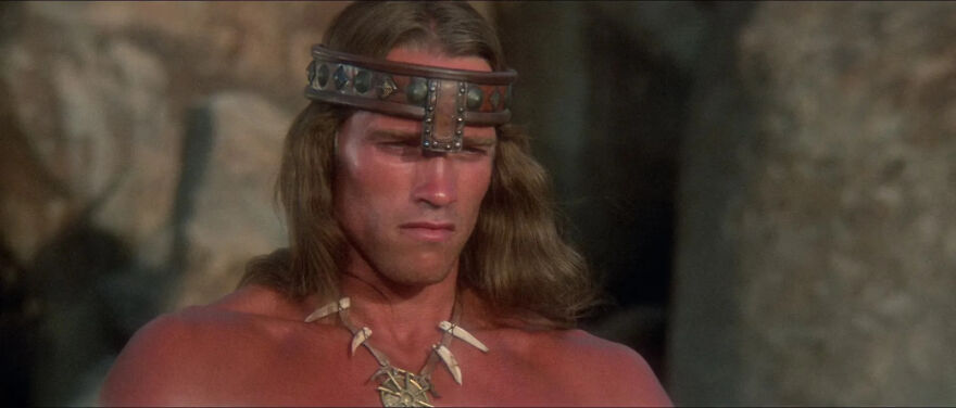 Actor Arnold Schwarzenegger in a warrior costume, wearing a headband and necklace, portraying a classic character.