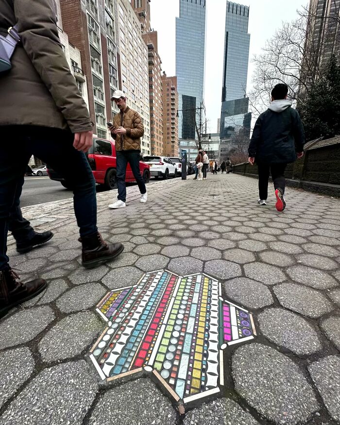 Vibrant mosaic art fills a cracked sidewalk in a city, surrounded by pedestrians and tall buildings.