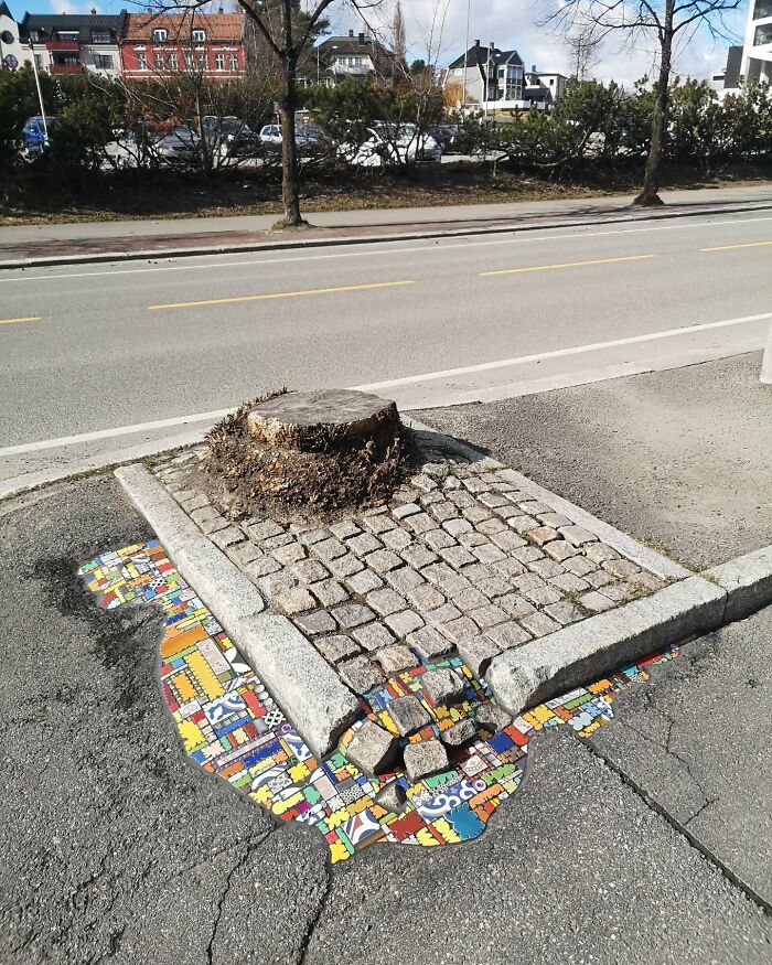 Colorful mosaic art fills a cracked sidewalk near a tree stump on a city street.