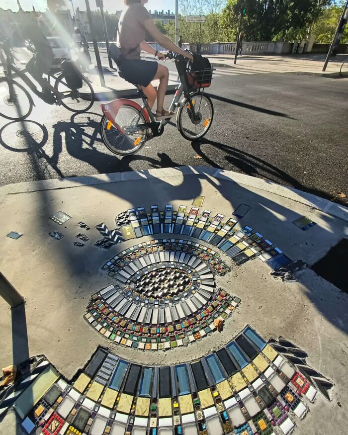 Vibrant mosaic artwork on sidewalk, patching a crack next to a cyclist in an urban setting.