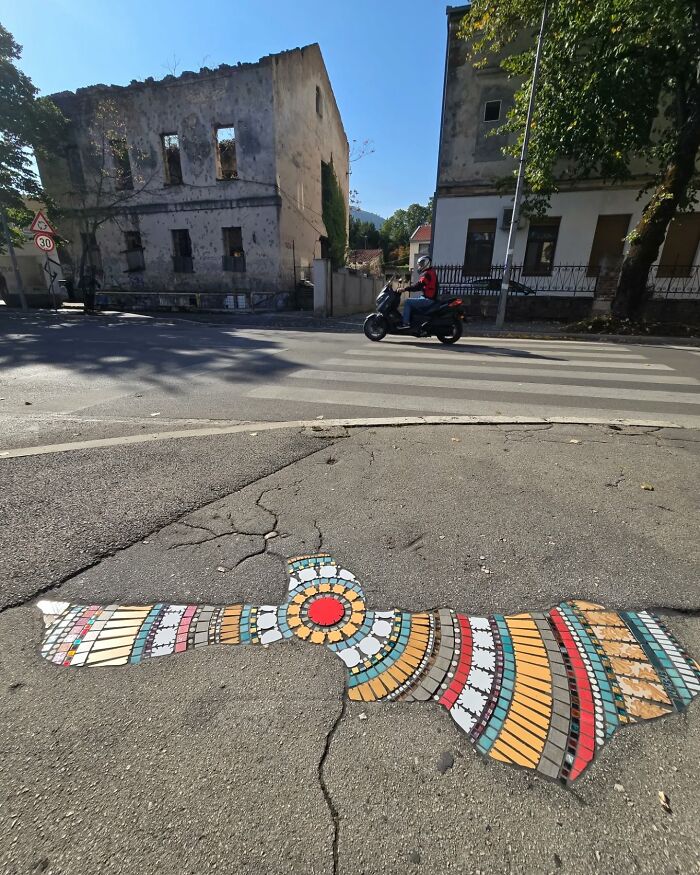 Vibrant mosaic art covering a cracked sidewalk, with old buildings and a motorcyclist in the background.