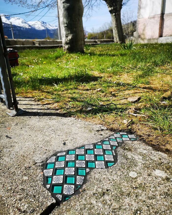 Vibrant mosaic art mends a cracked sidewalk near a grassy area with trees and mountains in the background.