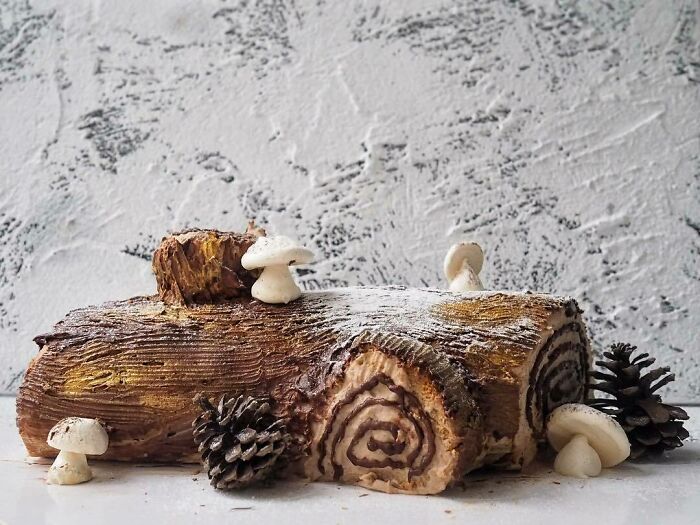 Festive yule log cake with chocolate swirl, dusted with powdered sugar, surrounded by pine cones and edible mushrooms; Christmas snack.