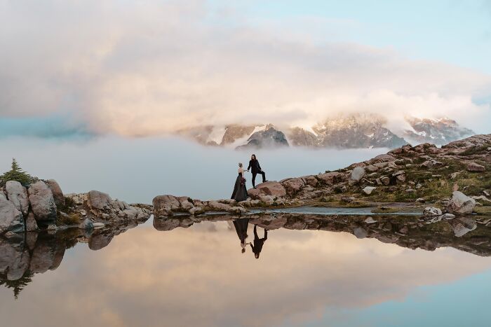 Mountain wedding photo with a couple walking on rocks by a reflective lake under a cloudy sky.