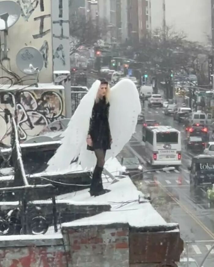 Person in angel costume standing on a snowy rooftop in New York City, surrounded by graffiti and traffic below.