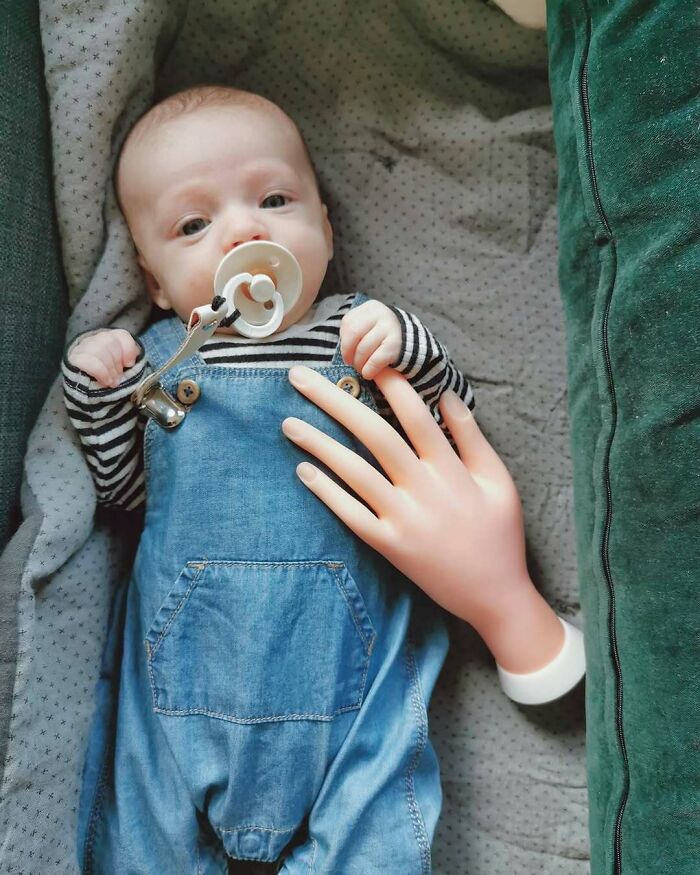 Baby in blue overalls with a pacifier and a toy hand, showcasing parenting tricks.