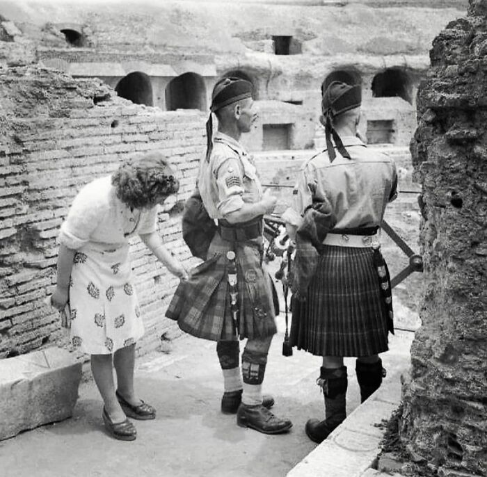 A woman examines the kilts of two soldiers at a historical site, reflecting on past traditions to shape future perspectives.