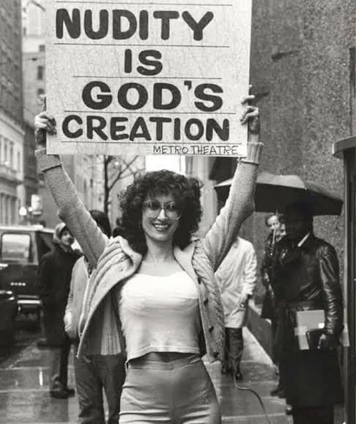 Woman holding a sign about nudity in a historical street scene, illustrating learning from the past to shape the future.