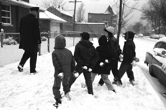 Children with snowballs follow a police officer in a historical winter scene on a snowy street.