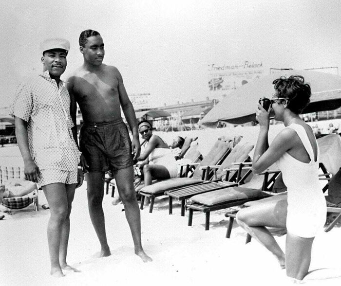 Vintage beach scene with two men posing while a woman takes a photo, capturing historical moments.