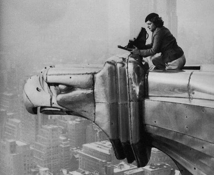 Woman perched on Chrysler Building gargoyle, capturing historical photos of New York City, embodying learning from the past.