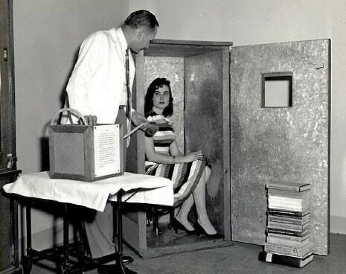 Man demonstrating an early metal soundproof booth with a woman seated inside, historical picture showcasing past technology.