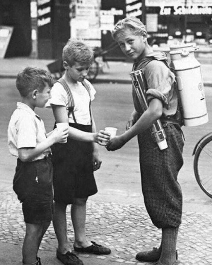 Three boys in vintage clothing share a drink on a street, capturing a historical moment.