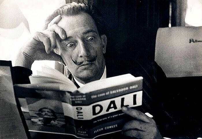 A person with a unique mustache intently reading a book by Salvador Dalí, reflecting on historical insights.