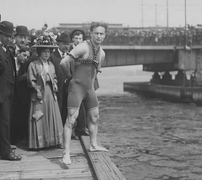 Man in chains on a dock, preparing for a stunt, surrounded by onlookers; historical picture.