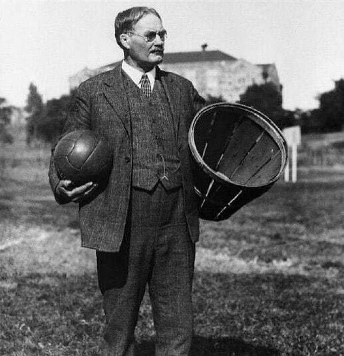 Man in a suit holding a vintage basketball and basket, showcasing historical sports equipment in an outdoor setting.