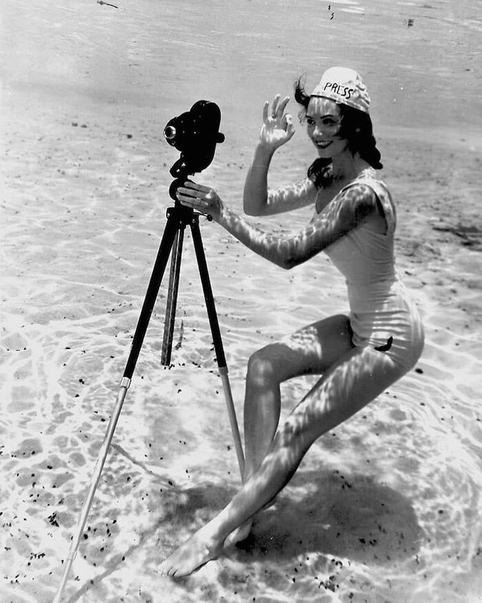 A woman in a swimsuit, wearing a press cap, operates a camera underwater, illustrating historical photography techniques.