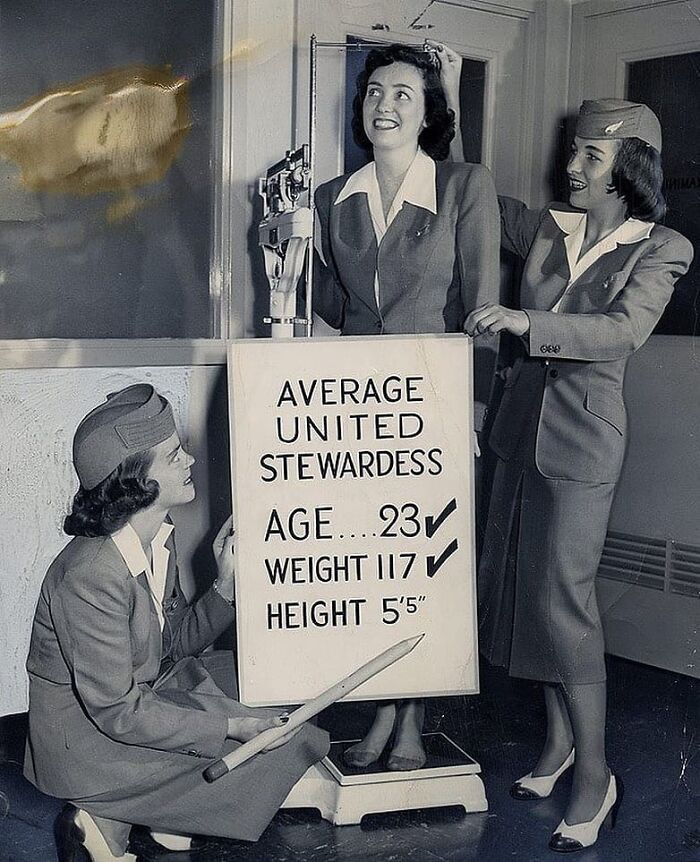 Vintage photo of stewardesses with a height and weight measurement board, illustrating historical airline standards.