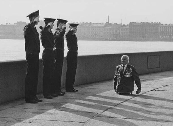 Veteran with medals saluted by young officers near waterfront, highlighting the impact of the past on the future.