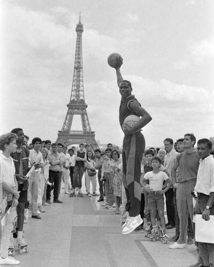 Basketball player posing mid-air with Eiffel Tower in background, surrounded by onlookers, showcasing historical moments.