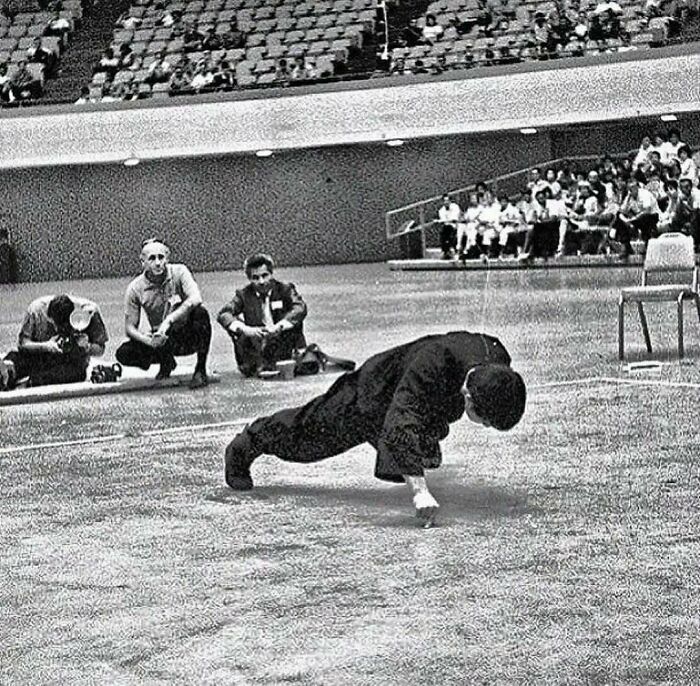 Martial artist performs one-arm push-up at a historical event, with spectators in the background.