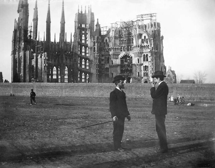 Two men in hats converse near the historical construction site of the Sagrada Familia, showcasing the past's architectural development.