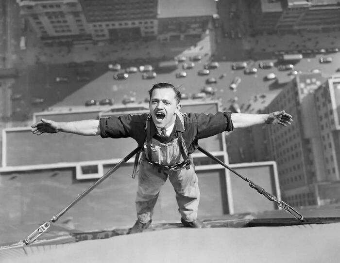 Man balanced on skyscraper edge in historical photo, arms outstretched, overlooking city traffic below.