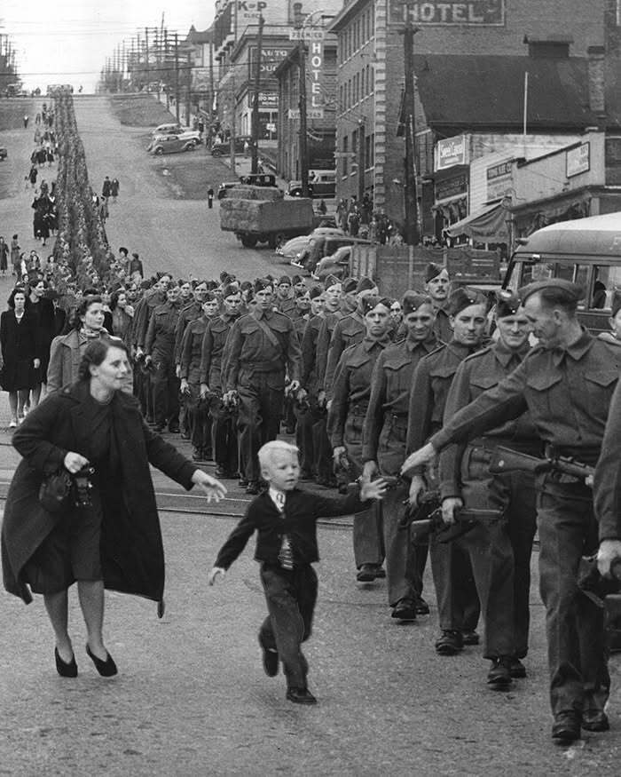 Historical scene of a woman reaching for a child running towards soldiers marching in a city street.