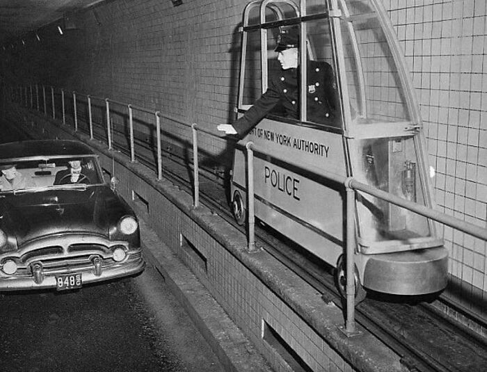 Police officer in a mobile booth inside a tunnel, New York, alongside a vintage car. Historic transportation scene.